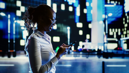 Joyful woman discussing with her friends using smartphone while wandering around dimly illuminated city streets at night. Chipper girl walking outside in urban environment, texting people