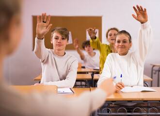 Teenage boys and girls sitting at desks in classroom and raising hands to answer question. Teacher...