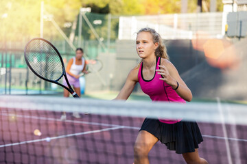 Young forcefully teenage girl playing tennis close to net on court outdoors