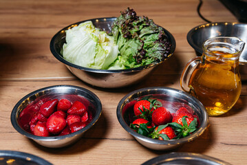 Bowls with raw cooking ingredients, a bowl of fresh strawberries, a bowl of lettuce leaves, a pitcher of oil on a wooden table surface