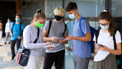 Teen students in medical face masks standing with workbooks in schoolyard during break in lessons. Concept of back to school after lockdown