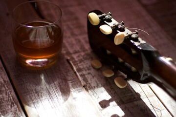 headstock of an acoustic guitar next to a glass with alcohol, on aged and darkened white wood.