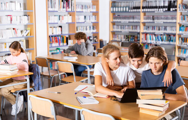 Three schoolchildren preparing for lessons in the school library are looking for the necessary information on a laptop, ..discussing topical issues