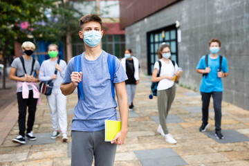 Confident teen boy in medical mask walking outside school building on autumn day, going to lessons....