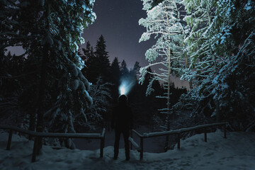 Night scene, a man with a headlamp in the winter forest by the frozen river. Starry sky.