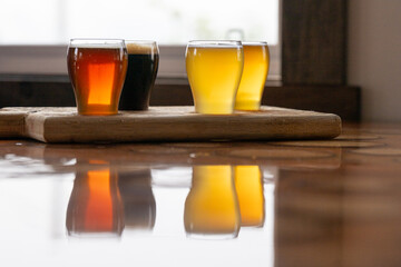 Small curved tulip shaped sample glasses of various ale beers with foam on the top. The samples are stout, red ale, pale ale, and IPA style beer on a wooden flight tray. The samples are on a table.