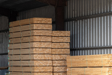 Wood boards stocked in a lumber yard. The two-by-four timber pieces are stacked high in a metal warehouse for consumer purchase. The construction materials are piled high from a carpentry sawmill.