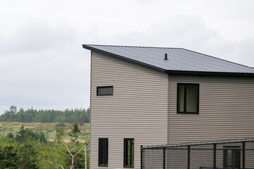 The exterior corner of a modern wooden tiny house. It has grey vinyl walls with black trim, closed glass windows, slanted flat roof, black metal backyard fence and lush green views of the countryside.