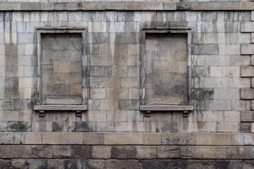 The exterior of an old brick building with two window frames sealed with grey mason blocks and concrete. The weathered warehouse or factory wall is dirty. The distressed beige brick material is rough.