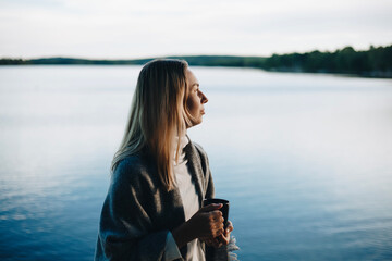 Beautiful young woman relaxing over a cup of tea. A beautiful blonde woman drinks a hot drink from a dark mug in an autumn park close to lake on the sunset. The woman is wrapped in a warm grey scarf.
