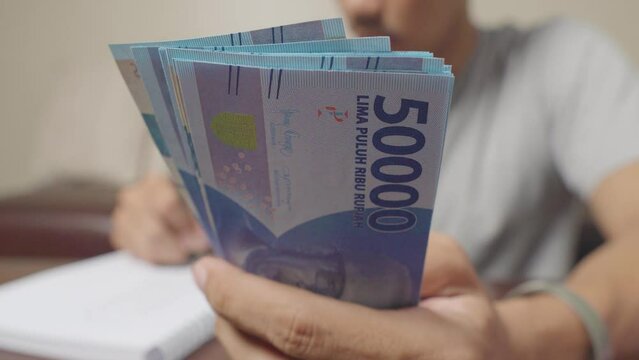 Close up of man's hand holding Indonesian rupiah banknotes and making notes, money financial management concept. Soft focus, Selective focus