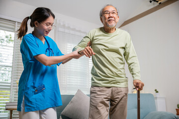 International Day for the Elderly. Young carer supporting helping senior man stand up with walking stick from sofa, Nurse wearing blue uniform help helping his patient senior old man with walking cane - obrazy, fototapety, plakaty
