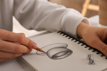Woman drawing cup of drink with graphite pencil in sketchbook at table, closeup