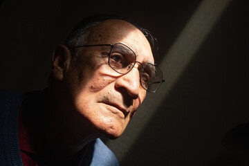 Old man or elder model with glasses with his face illuminated in the dark, black background. Caucasian or latino wearing blue clothes