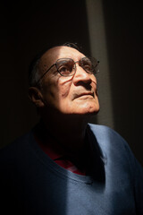 Old man or elder model with glasses with his face illuminated in the dark, black background. Caucasian or latino wearing blue clothes