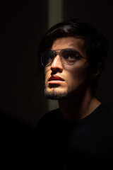 Portrait of a young handsome male model with glasses and beard with his face illuminated in the dark, black background. Caucasian or latino.