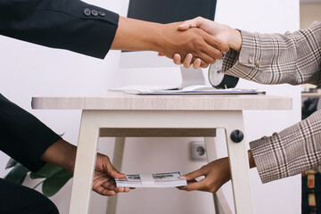 Two business people shaking hand and taking bribe money under wooden working table at the office. 