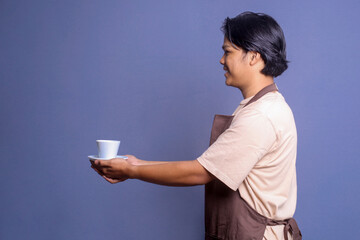 Side view of young barista guy wear brown apron holding and serving cup of coffee against blue background