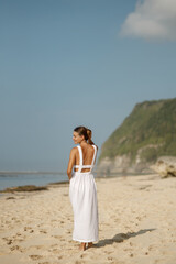 Fototapeta na wymiar Back view of beautiful brunette girl in a white linen dress walking by seaside. Travel vacation relaxation abroad lifestyle
