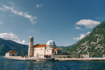 Group of tourists stands on the island of Gospa od Skrpjela near a moored boat. Montenegro - obrazy, fototapety, plakaty