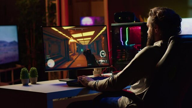 Man in dark living room playing videogame on gaming PC at computer desk, relaxing by shooting enemies. Gamer battling flying robots in online singleplayer shooter from neon lights ornate home