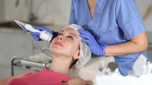 Female cosmetologist performing anti-aging face phototherapy procedure, shrink pores and repair damaged skin for young girl client in clinic of aesthetic medicine