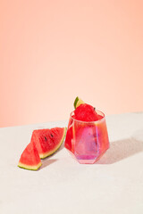 Fresh watermelon is placed next to a glass of juice on a white surface with a pastel pink background. The antioxidants found in watermelon provide many health benefits.