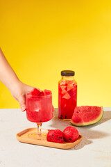A hand is placing a glass of watermelon juice on a wooden tray, next to a bottle of detox water and fresh watermelon. White surface with yellow background.