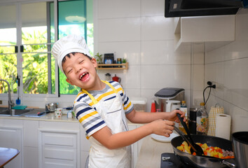 Cute happy smiling Asian 8 years old boy child wearing chef hat and apron having fun preparing,...
