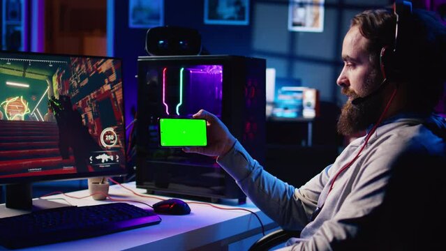 Man watching tutorial on mockup phone while playing first person shooter videogame with gun shooting laser bullets. Gamer learning to play game by looking at online guide on chroma key device