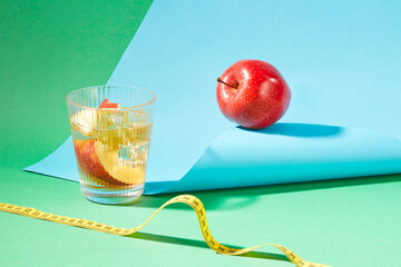 A glass of detox water with an apple, a tape measure and a blue sheet of paper on a green background. Apples are rich in nutrients, fiber and low in calories to help support weight loss.