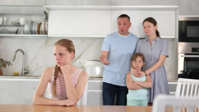  At home kitchen, parents scold daughter for careless act. Sulky preschool girl sit near table during serious conversation with parents