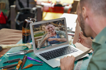 Closeup and back view of leather craftsman using a laptop video call to customer to show a color of...