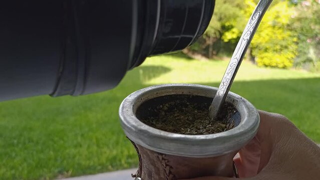 Pouring hot water from thermos into Mate while holding it in hand, traditional drink from Argentina, close up