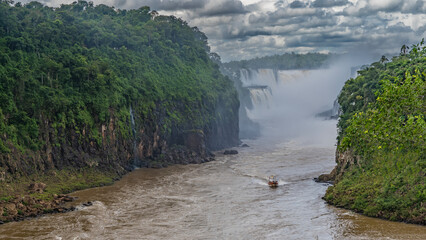 A tourist boat rushes along a stormy river. The cascades of the waterfall are shrouded in spray and...