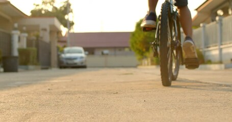 Slow motion low angle shot while a 9 year old Asian boy rides a bicycle in the evening on a weekend in an urban village and there is a beautiful golden sunlight in the scene. - Powered by Adobe