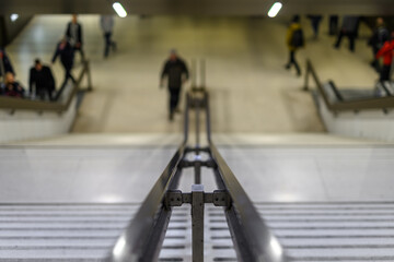 A stainless steel railing of a pedestrian staircase into the railway with blurred people in the...