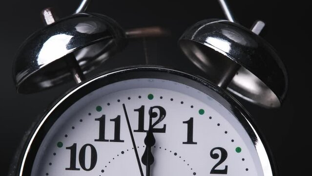 The old alarm clock was ringing at 12:00 AM or 12:00 PM. Close-up of a retro alarm clock face in time lapse on black background. Clock hands move fast.