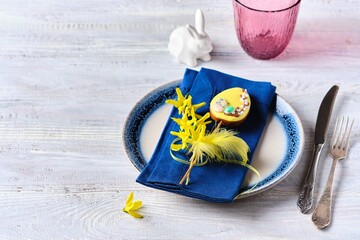 Easter table setting with blue napkins, burgundy water glasses and yellow decor on a wooden table. Easter lunch concept.