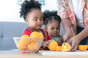 Little African cute daughters helping her senior father to slice orange on cutting board and...