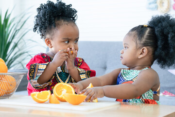 African cute kid girls have fun and enjoy eating fresh orange together for breakfast at home. Two siblings eating sliced orange in hands with messy mouth. Healthy lifestyle and learning concept