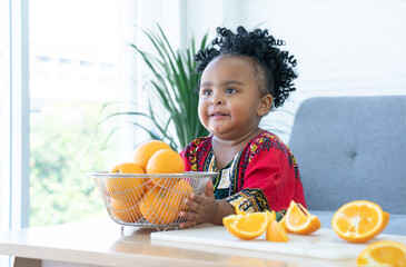 African cute kid girl try to lift fresh oranges basket up at home. Adorable child eating sliced...