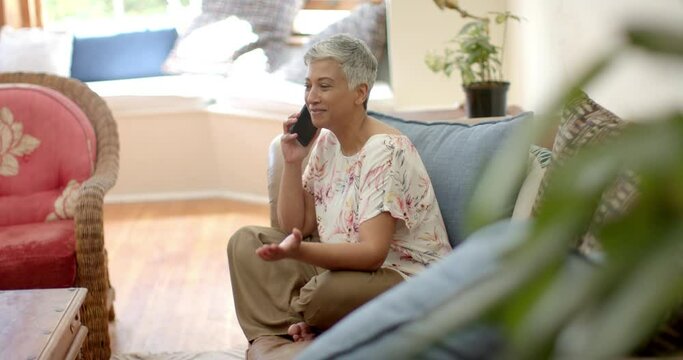 Happy senior biracial woman sitting on couch and talking on smartphone at home, slow motion