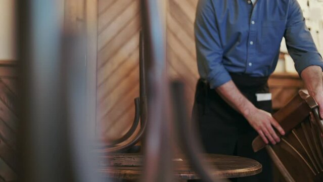 Cropped of male waiter dressed in uniform arranging wooden chairs near tables while opening retro style restaurant in morning. Caucasian man following service demands for clients at start of shift.