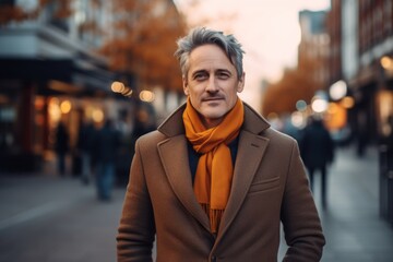 Handsome middle-aged man with gray hair wearing a brown coat and orange scarf on a city street.