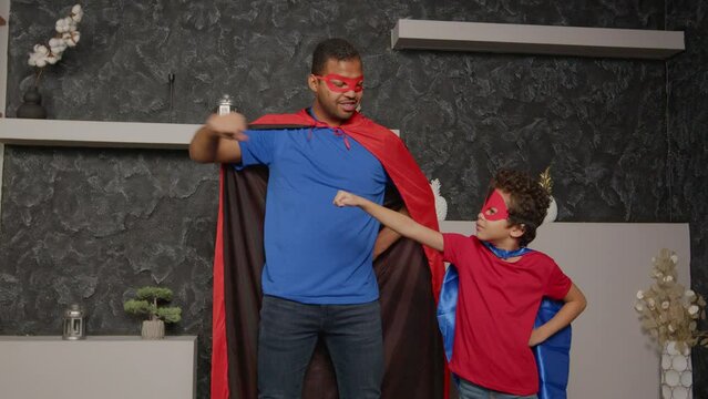 Confident attractive African American father and adorable school age son in superheroes costumes standing in flying pose, making fist bump gesture, showing power ,strength and togetherness at home.