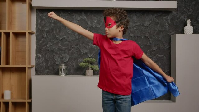 Portrait of determined cute school age boy wearing superhero cape and mask, standing in flying pose and running away while carefree child having playing and imaging to be hero indoors.