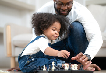 Cute little kid playing chess board toy as her parent sitting by. Multiracial family joyful game...