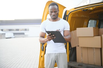african american Worker unloading boxes from van outdoors. Moving service concept. Male employee of moving company makes inventory of packed boxes and writes data to clipboard.