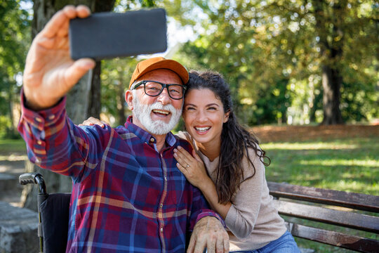 Smiling senior man in wheelchair taking selfie with caregiver at public park